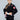 A young girl with long brown hair in a ponytail stands confidently wearing the Kids Competition BJJ Gi Black, featuring red and white patches, and holding a white belt against a plain gray background.