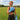 Woman wearing black athletic wear and blue boxing gloves on a grassy field with trees and buildings in the background.